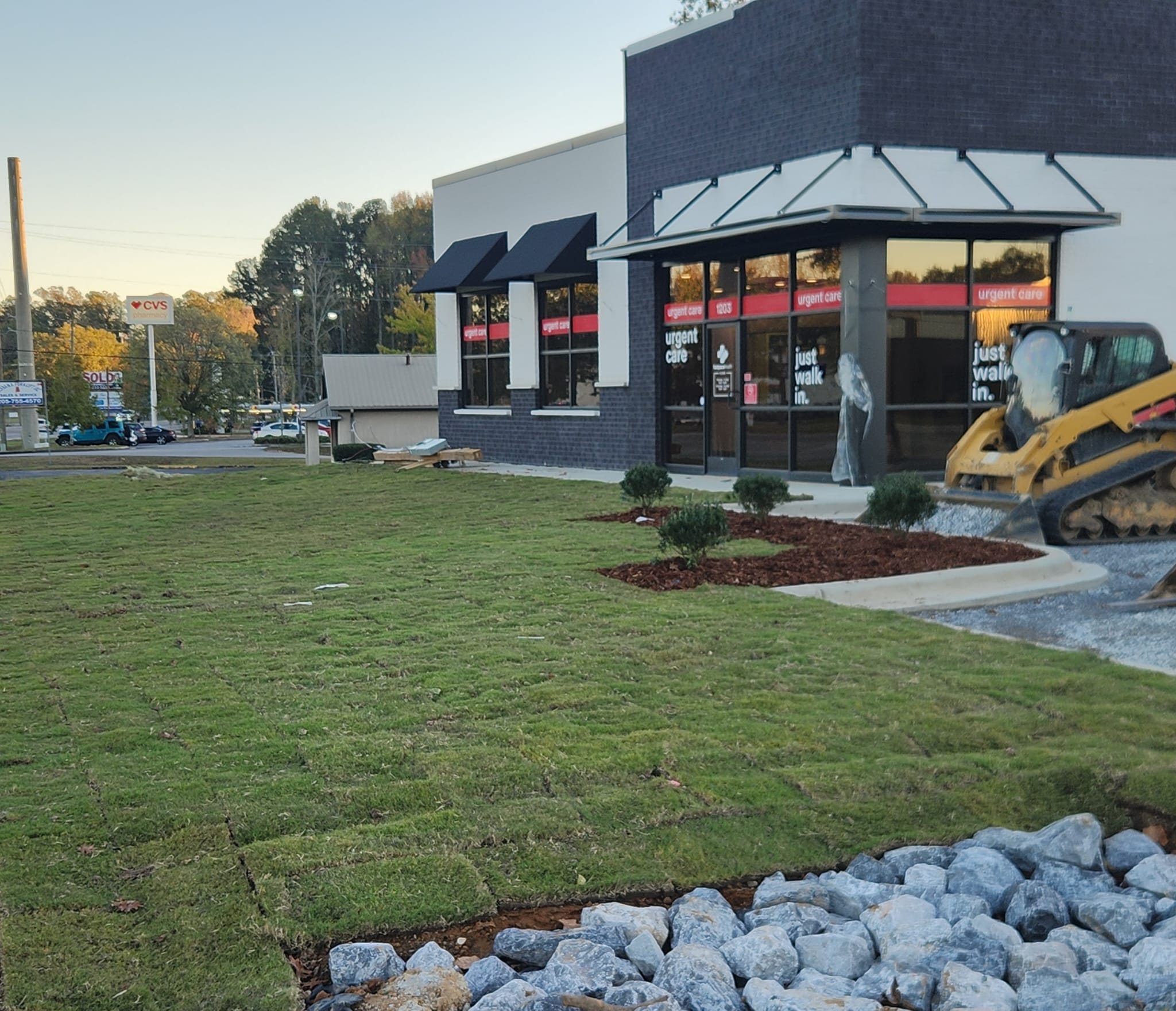 Freshly installed sod and rock border in front of a commercial storefront, with a skid steer on site
