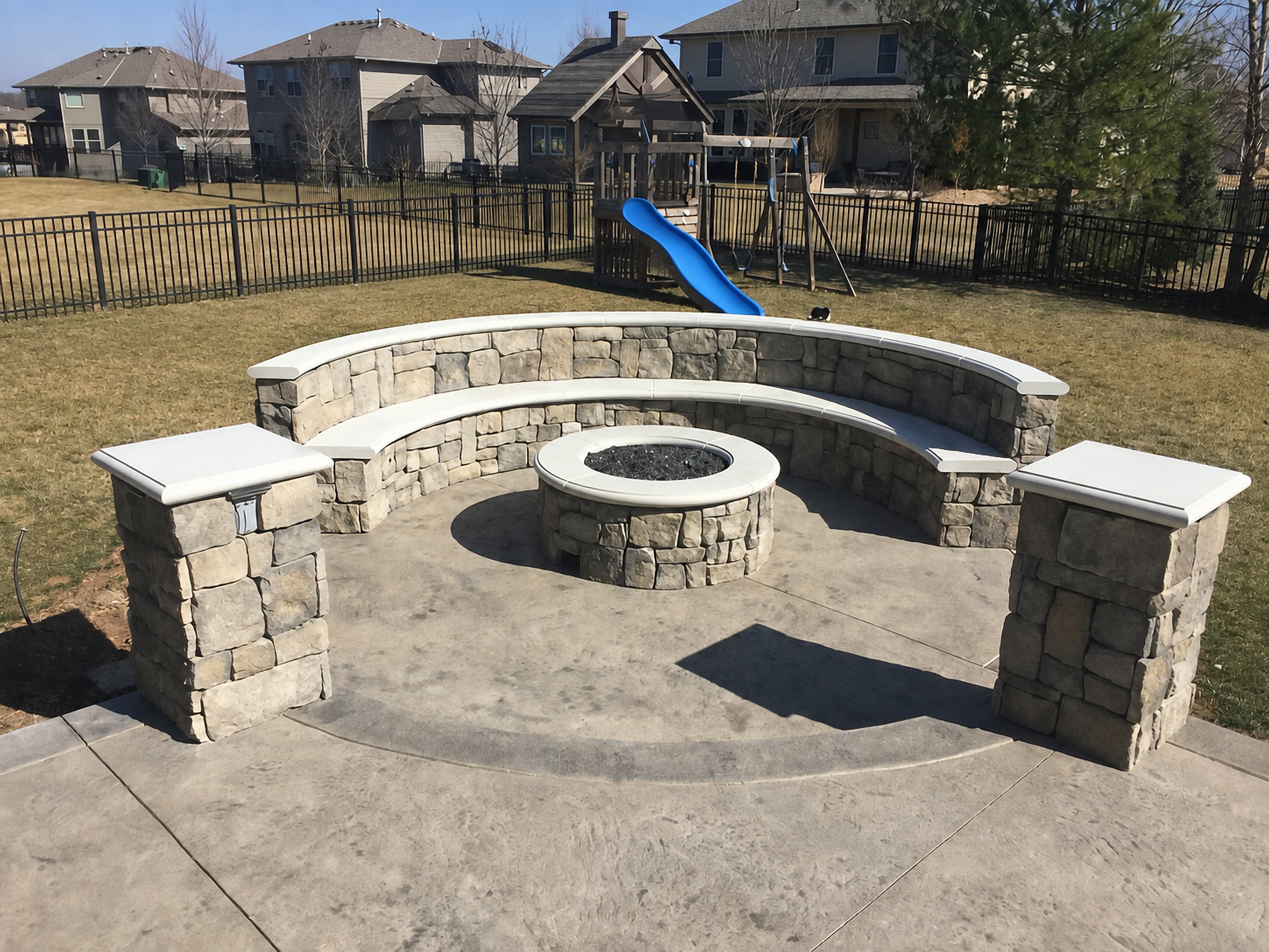 Fire pit with curved seating wall, white capstones, and stone pillars on a stamped concrete patio