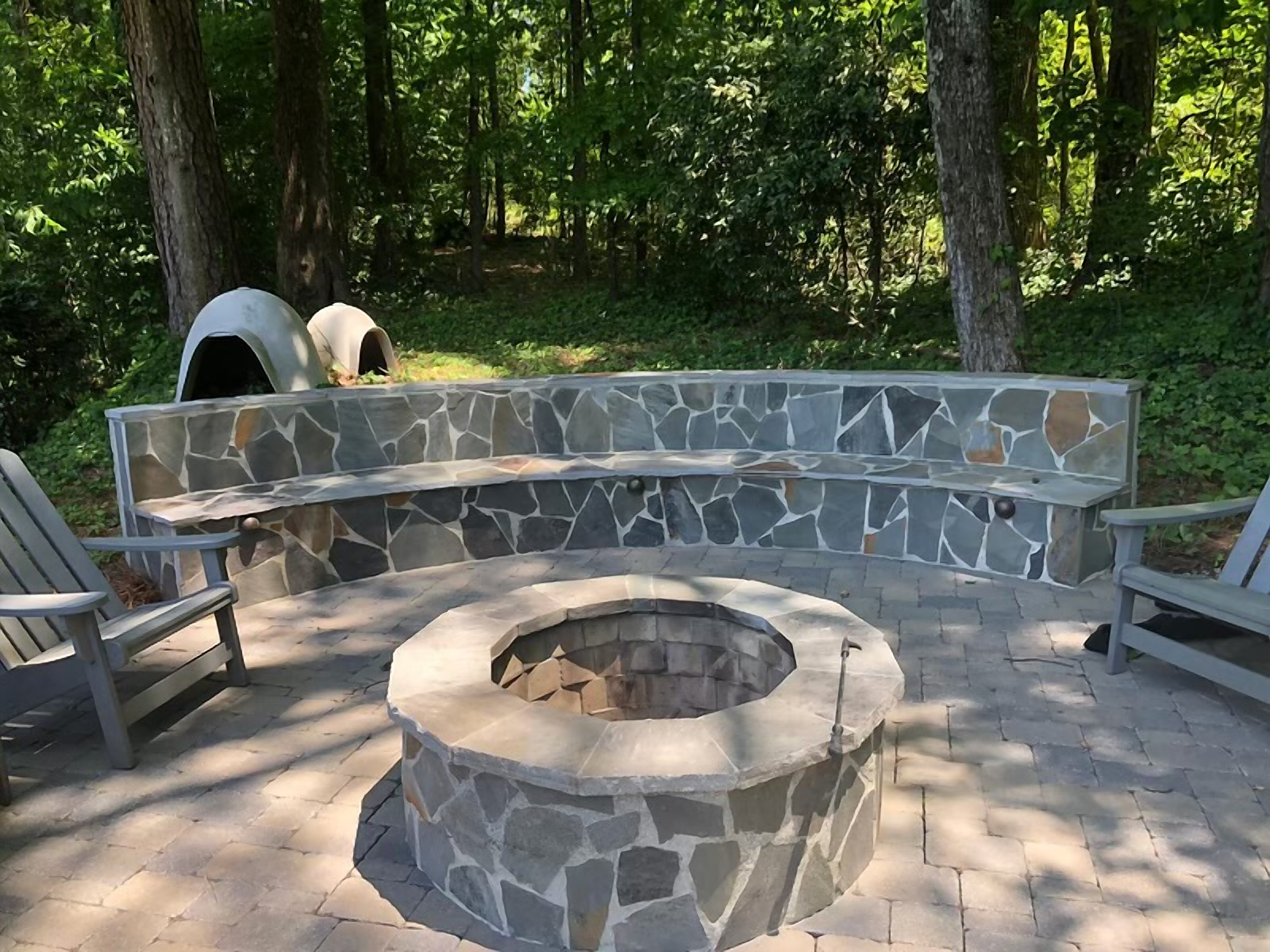 Stone fire pit and curved seating wall on a paver patio surrounded by mature trees